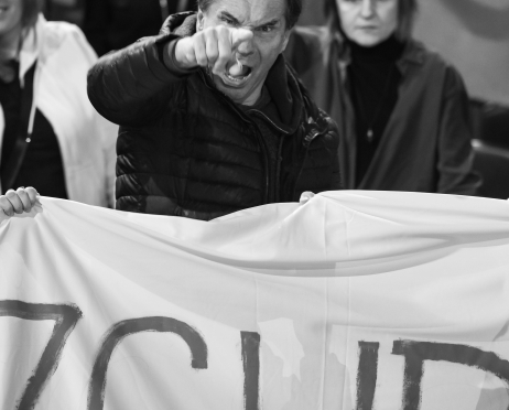 Czarno-białe zdjęcie. Aktor Wojciech Skibiński stoi wśród protestujących w teatrze. Na pierwszym planie trzyma duży baner i z wyraźną złością wskazuje palcem przed siebie. Za nim stoją inne osoby, część z uniesionymi rękami, w tle widoczna jest polska flaga.